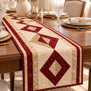 May include: A rectangular fabric table runner featuring a geometric quilt pattern with alternating deep red and cream-colored diamond shapes. The runner has intricate stitched leaf motifs and floral borders in shades of red and tan.