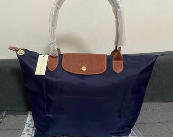 Op de afbeelding: Een marineblauwe nylon tas met een bruine leren klep en een goudkleurige drukknoopsluiting. De tas heeft twee handvatten omwikkeld met wit beschermpapier en een crèmekleurig label met de tekst LONGCHAMP eraan bevestigd.