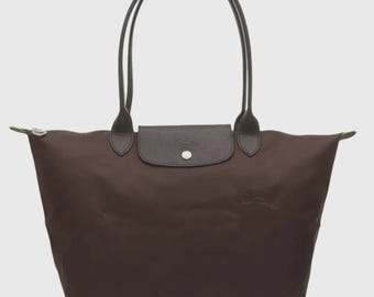 Pode incluir: Uma bolsa tote de nylon castanho-escuro com duas alças longas, uma aba de couro texturizado com fecho de botão de pressão prateado e um logótipo subtilmente gravado na frente. A bolsa tem uma forma trapezoidal larga com fecho de correr.
