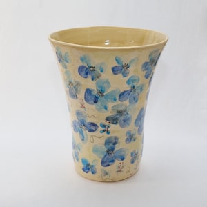 May include: A ceramic vase with a light yellow glaze and a blue floral pattern. The vase has a flared opening and a rounded bottom.