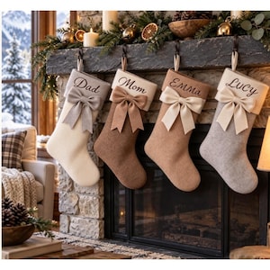 Peut inclure: Quatre chaussettes de Noël personnalisées suspendues en rangée. Les chaussettes sont crème, beige, marron clair et gris, chacune avec un grand nœud en tissu et des noms brodés : Dad, Mom, Emma et Lucy. Elles sont en feutrine douce.