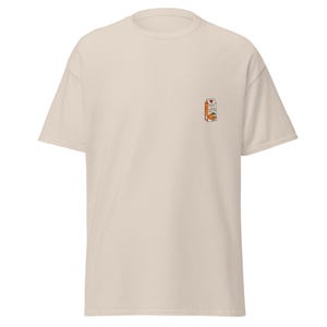May include: A beige short-sleeve t-shirt featuring a small graphic on the upper left chest. The graphic depicts a miniature orange juice carton with the text "ORANGE JUICE SOME BUGS" printed on the front of the carton, accompanied by an orange.