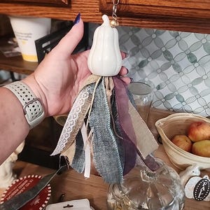 May include: A white ceramic pumpkin ornament attached to a metal lobster clasp and a gold bead, featuring a decorative tassel made of layered fabric strips in shades of tan, blue, and dark purple, including lace and burlap textures.
