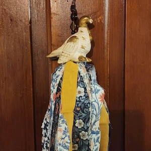 Può includere: Un ornamento decorativo da appendere con una statuetta di uccello color crema e oro posizionata sopra un gruppo di strisce di tessuto. Il tessuto presenta motivi floreali in blu navy, bianco e arancione, accentuati da nastri giallo senape e perline scure.