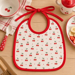 Puede incluir: Un babero de tela blanca para bebé con un patrón repetitivo de pequeñas cerezas rojas y diminutos puntos rojos. El babero tiene un borde de tela roja brillante y un lazo de tela roja a juego en la parte superior para atarlo al cuello.