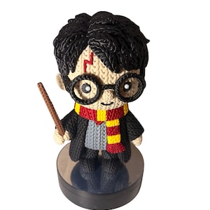 Puede incluir: Una pequeña figura de un niño con cabello negro, gafas redondas negras y una cicatriz roja en forma de rayo en la frente. La figura viste un suéter gris, una túnica negra y una bufanda a rayas rojas y amarillas, sosteniendo una varita marrón.