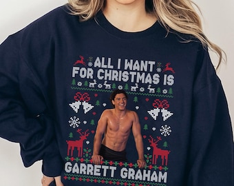 May include: A navy blue crewneck sweatshirt featuring a holiday-themed graphic with the text "ALL I WANT FOR CHRISTMAS IS GARRETT GRAHAM" surrounding a photographic print of a shirtless man, accented by red and white reindeer and snowflakes.