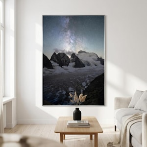 Puede incluir: Una impresión de arte mural vertical que muestra un paisaje nocturno de picos montañosos nevados bajo un cielo estrellado con la Vía Láctea visible. La escena incluye laderas rocosas oscuras y un glaciar, con un jarrón negro con hierba seca.