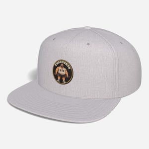 Puede incluir: Una gorra de béisbol de color gris claro jaspeado con visera plana y un parche circular en la parte delantera. El parche presenta una ilustración de dibujos animados de una figura musculosa sosteniendo mancuernas con el texto ENDOMORPH.