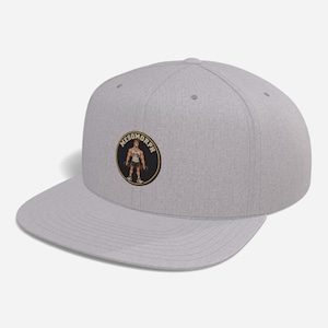 Puede incluir: Una gorra snapback de color gris jaspeado con un parche circular en la parte frontal. El parche tiene un fondo negro, un borde dorado y el texto MESOMORPH en dorado. Dentro del círculo hay una ilustración de una figura musculosa con mancuernas.