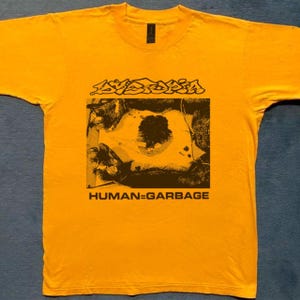 Peut inclure: Un t-shirt jaune vif à manches courtes avec une impression graphique noire sur la poitrine. Le motif comprend le texte "DYSTOPIA" au-dessus d'une illustration sombre d'un arbre et "HUMAN=GARBAGE" en lettres capitales noires sous l'image.
