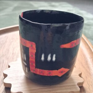 May include: A black ceramic cup with a glossy finish, featuring a hand-painted orange geometric band with small green dots and white vertical dash markings. The cup has a slightly irregular, organic rim and sits on a light-coloured wooden coaster.