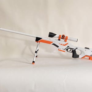 Puede incluir: Un rifle de francotirador de juguete en blanco, naranja y negro con un cañón largo, una mira telescópica con detalles naranjas y un bípode plegable. El cuerpo presenta patrones geométricos y detalles negros, con una culata naranja.