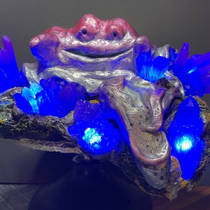 Könnte beinhalten: Eine skurrile Skulptur einer rosa-silbernen Kreatur, umgeben von blauen Kristallen. Die Kreatur hat ein breites Lächeln und sitzt auf einem dunkelbraunen Felsen. Die Kristalle leuchten blau und erzeugen einen magischen Effekt.
