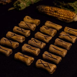 May include: Wooden rune stones with etched symbols, arranged on a dark surface. The runes are light brown with dark brown markings. A cob of corn and herbs are in the background.