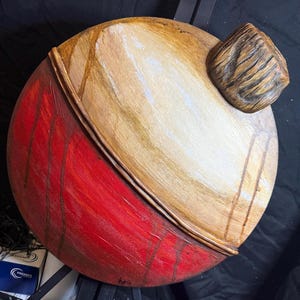 May include: A round, wooden, red and brown painted buoy with a wooden top. The buoy is painted with red and brown stripes and has a white stripe around the middle. The buoy is hanging from a black hook.