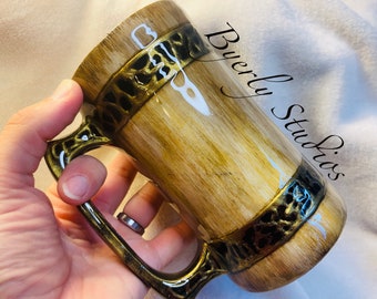Könnte beinhalten: Eine braune und goldene Keramiktasse mit Holzmaserung und einem schwarzen und goldenen Musterband um den oberen Rand. Die Tasse hat einen Henkel mit einem ähnlichen Muster. Die Tasse ist mit "Byerly Studios" beschriftet.