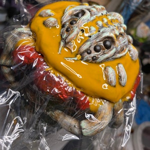 May include: A ceramic sculpture of a creature with a yellow glaze, green and brown details, and black eyes. The sculpture is covered in plastic wrap.