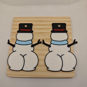 Könnte beinhalten: Ein Paar schneemannförmige Ohrringanhänger mit weißem Körper, schwarzem Zylinder mit rotem Band und hellblauem Schal. Jeder Schneemann hat schwarze Stockarme und einen ausgeprägten, abgerundeten unteren Bereich. Die Anhänger liegen auf einem Holzbrett.