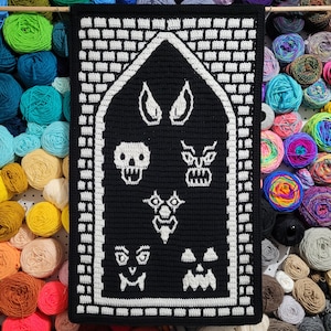 May include: A black and white crochet wall hanging featuring various spooky faces and a skull. The design is framed with a white brick pattern and a black border. The background shows colorful yarn skeins.