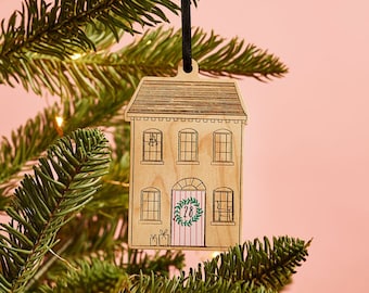 May include: A wooden Christmas ornament shaped like a house with a pink door and a wreath with the number 28 on it. The ornament is hanging from a black ribbon on a pine tree branch.