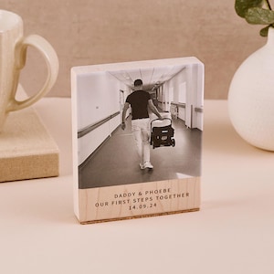 May include: A black and white photograph of a person pushing a baby stroller down a hospital corridor. The photo is printed on a wooden block with the text "Daddy & Phoebe Our First Steps Together 14.09.24" printed on the bottom.
