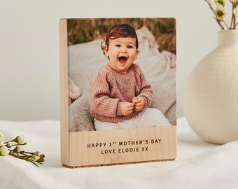 May include: A wooden block with a photograph of a baby wearing a pink sweater. The block has the text "HAPPY 1ST MOTHER'S DAY LOVE ELODIE XX" engraved on the bottom. A white vase with flowers is in the background.