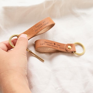 Op de afbeelding: Twee lichtbruine leren sleutelhangers met messing ringen en klinknagels. Eén sleutelhanger is gegraveerd met "Happy 3rd anniversary!" en de andere met "Hannah ♥ Sam 12th September 2026". Het leer is in een lus gevouwen en vastgezet met een metalen klinknagel.