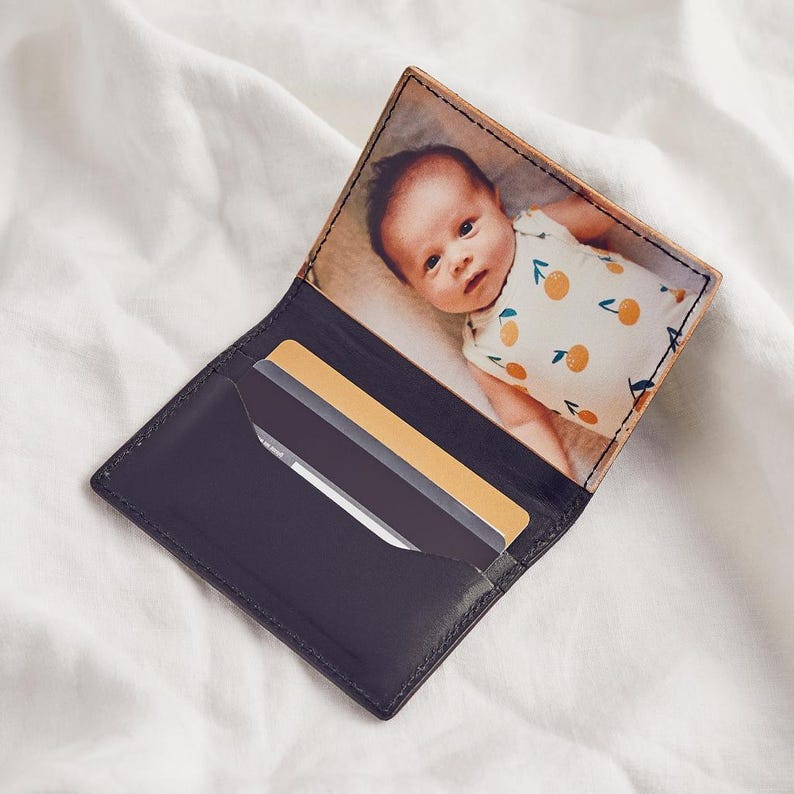 K&ouml;nnte beinhalten: Schwarzes Lederportemonnaie mit einem individuellen Foto eines Babys im Inneren. Das Portemonnaie hat ein Kartenfach mit zwei sichtbaren Karten. Das Baby tr&auml;gt einen wei&szlig;en Strampler mit orangefarbenen Fruchtmustern.