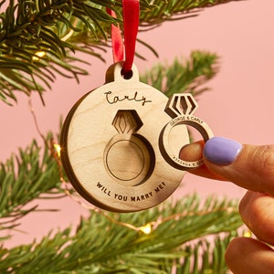 May include: A round wooden ornament hanging from a red ribbon, featuring a cutout in the shape of a diamond ring. The wood is engraved with the name Carly and the text Will You Marry Me? A matching wooden ring cutout displays George & Carly.