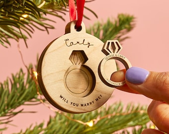 Op de afbeelding: Een ronde houten hanger aan een rood lint, met een uitsparing in de vorm van een verlovingsring. Het hout is gegraveerd met de naam Carly en de tekst Will You Marry Me? Een bijpassende houten ring toont de tekst George & Carly.