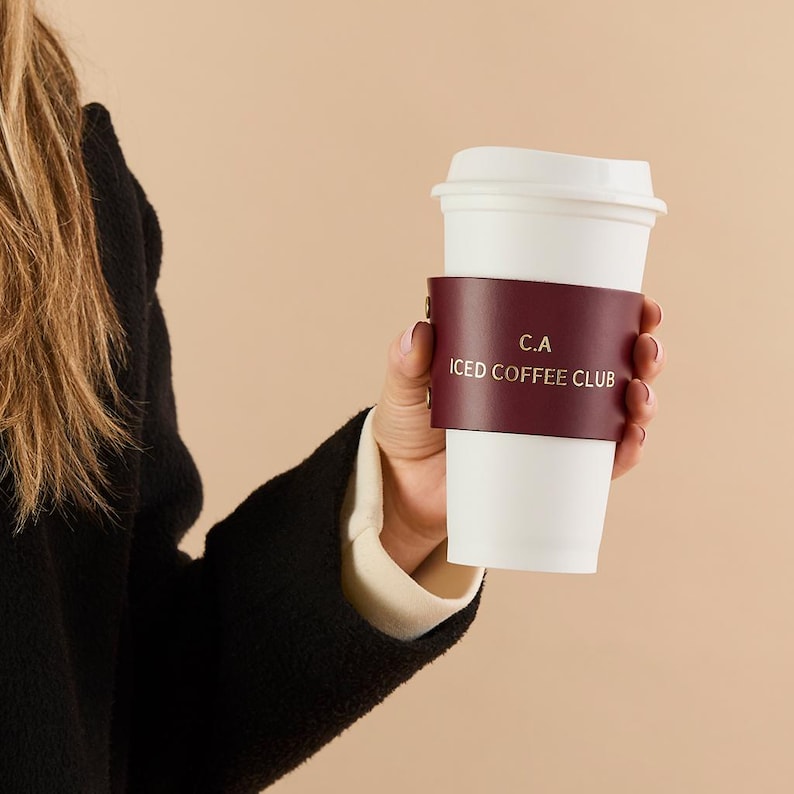 May include: A white paper coffee cup with a burgundy leather sleeve. The sleeve has the text "C.A ICED COFFEE CLUB" printed on it. The cup is held by a person wearing a black jacket.