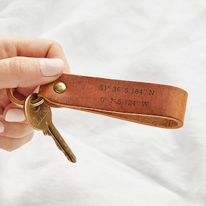 May include: A tan leather keyring with a brass key ring and a key. The keyring features the engraved coordinates "51°36'5.184" N 0°7'5.124" W". The leather is secured with a brass rivet.