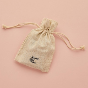 May include: A beige drawstring pouch with the words "create gift love" printed in black. The pouch is made of a textured fabric and is set against a pink background. The drawstring is tied at the top.