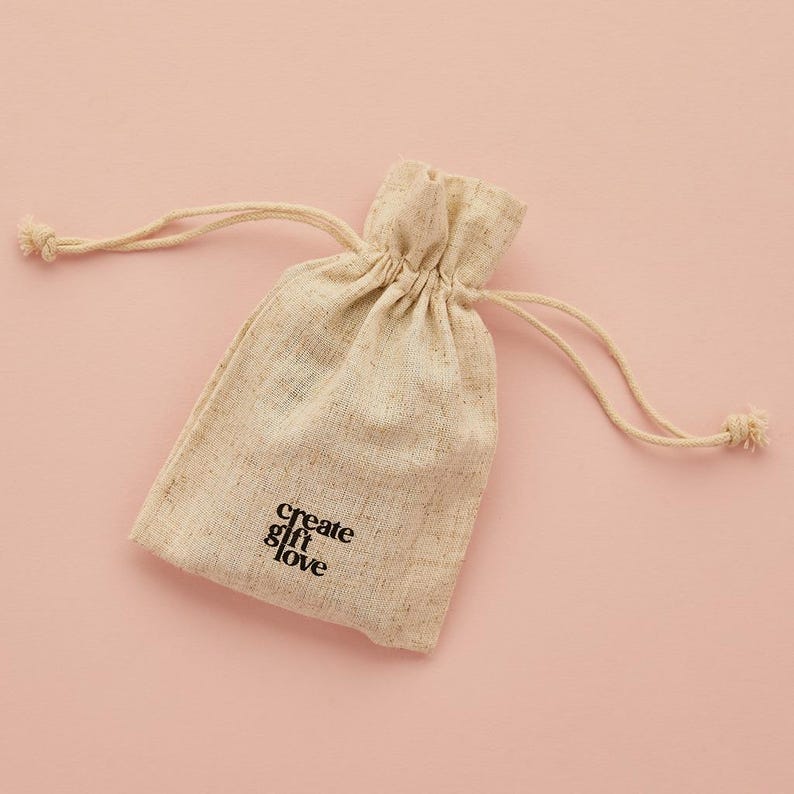 May include: A beige drawstring pouch with the words "create gift love" printed in black. The pouch is made of a textured fabric and is set against a pink background. The drawstring is tied at the top.