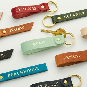 Op de afbeelding: Een collectie leren sleutelhangers in diverse kleuren, waaronder rood, olijfgroen, bruin, mintgroen, donkerbruin, blauwgroen en marineblauw. Elke sleutelhanger heeft een goudkleurige metalen ring, een gouden klinknagel en goudkleurige tekst zoals 22.07.2023, DADDY, SOPHIE, EXPLORE, BEACHHOUSE en MY PLACE.