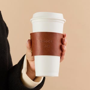 May include: A white paper coffee cup with a brown leather sleeve. The sleeve is embossed with the words "DADDY'S TEA". The cup is held by a person.