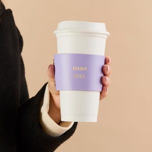 May include: A white paper coffee cup with a light purple leather sleeve. The sleeve has the words "MAMA FUEL" in gold lettering. The cup is held by a person.