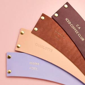 May include: A collection of leather drink sleeves in various colors, including purple, tan, and brown. Each sleeve has gold-colored rivets and embossed text such as "Mama Fuel," "Daddy's Tea," and "Iced Coffee Club."