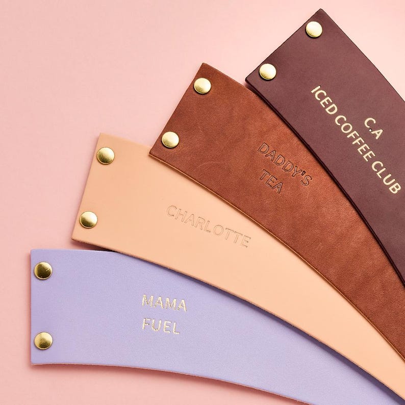 May include: A collection of leather drink sleeves in various colors, including purple, tan, and brown. Each sleeve has gold-colored rivets and embossed text such as "Mama Fuel," "Daddy's Tea," and "Iced Coffee Club."