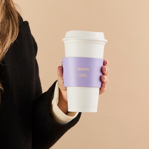 May include: A white paper coffee cup with a lid, held by a person. The cup has a light purple sleeve with the words "MAMA FUEL" in gold lettering. The person is wearing a black coat.