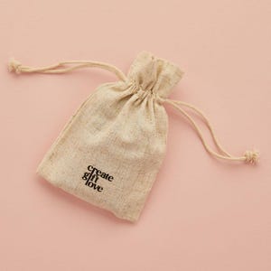 May include: A beige drawstring bag with the words "create & love" printed in black. The bag is made of a textured fabric and has a drawstring closure. The background is a solid light pink.