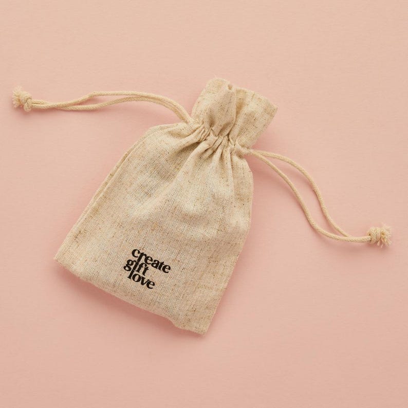 May include: A beige drawstring bag with the words "create & love" printed in black. The bag is made of a textured fabric and has a drawstring closure. The background is a solid light pink.