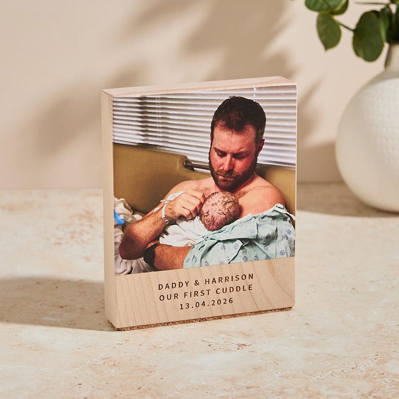 May include: A wooden block with a photograph of a man holding a newborn baby. The block has the text "DADDY & HARRISON OUR FIRST CUDDLE 13.04.2026" printed on the front. The image is in color.