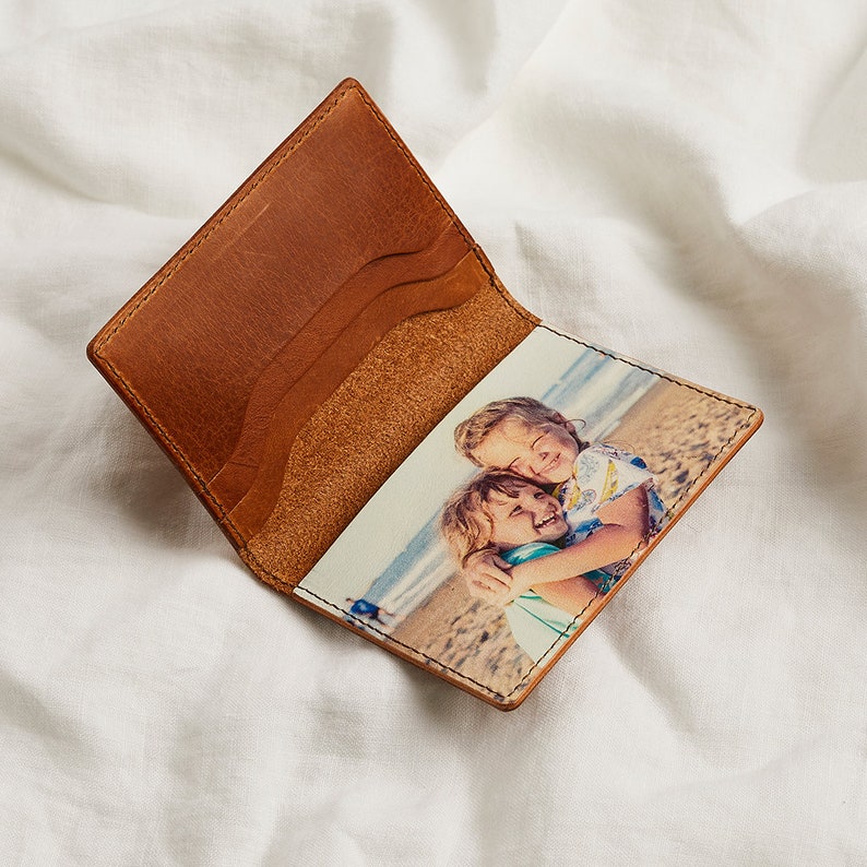 K&ouml;nnte beinhalten: Offene Ledergeldb&ouml;rse mit Fotoaufdruck. Die Geldb&ouml;rse ist ge&ouml;ffnet und zeigt ein gedrucktes Bild von zwei Kindern, die sich an einem Strand umarmen. Die Au&szlig;enseite ist hellbraun, mit mehreren Kartenf&auml;chern. Die Geldb&ouml;rse ist aus Leder.