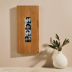 May include: A rectangular wooden photo frame with four black and white portrait photos. The frame is made of light brown wood and is mounted on a beige wall. A white vase with green leaves sits on a table below.