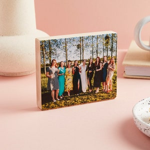Peut inclure: Un bloc de bois avec une photo de groupe de femmes en robes, posant dans un champ avec des arbres en arrière-plan. Le bloc rectangulaire est posé sur une surface rose. Un vase blanc et une tasse sont visibles.