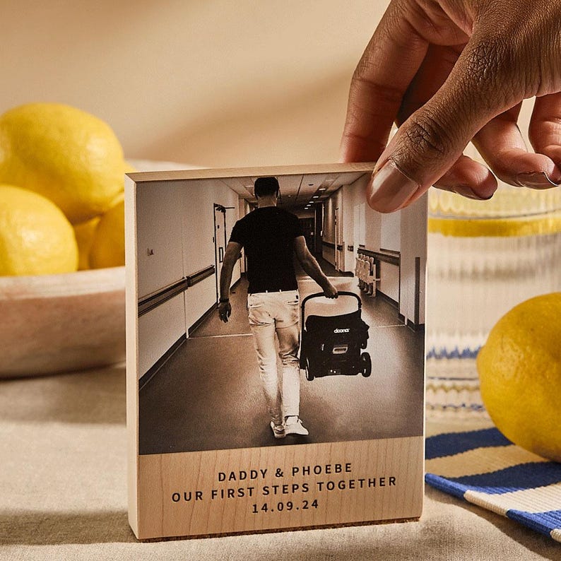 May include: A wooden block with a sepia-toned photograph of a person pushing a stroller down a hallway. The text "DADDY & PHOEBE OUR FIRST STEPS TOGETHER 14.09.24" is printed on the bottom of the block.