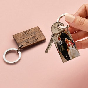 May include: A rectangular wooden keychain engraved with an August 2024 calendar and the text The day we tied the knot, paired with a second rectangular keychain featuring a printed photograph of a couple in wedding attire, both on metal rings.