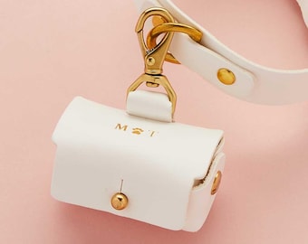 May include: A white leather-like waste bag dispenser with a gold-toned metal snap closure and a gold-toned swivel clasp. The front of the rectangular pouch features the gold-stamped text M, a paw print icon, and the letter T. It attaches to a matching collar.
